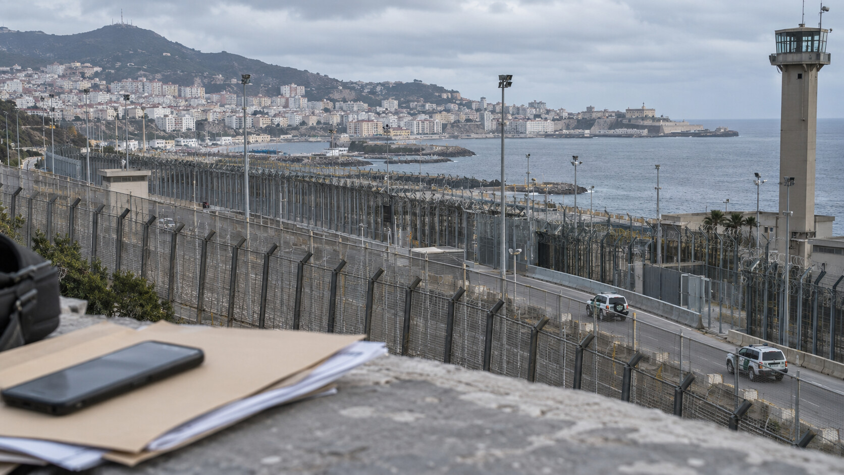Grenzanlagen von Ceuta mit Aktenmappe und Mobiltelefon im Vordergrund