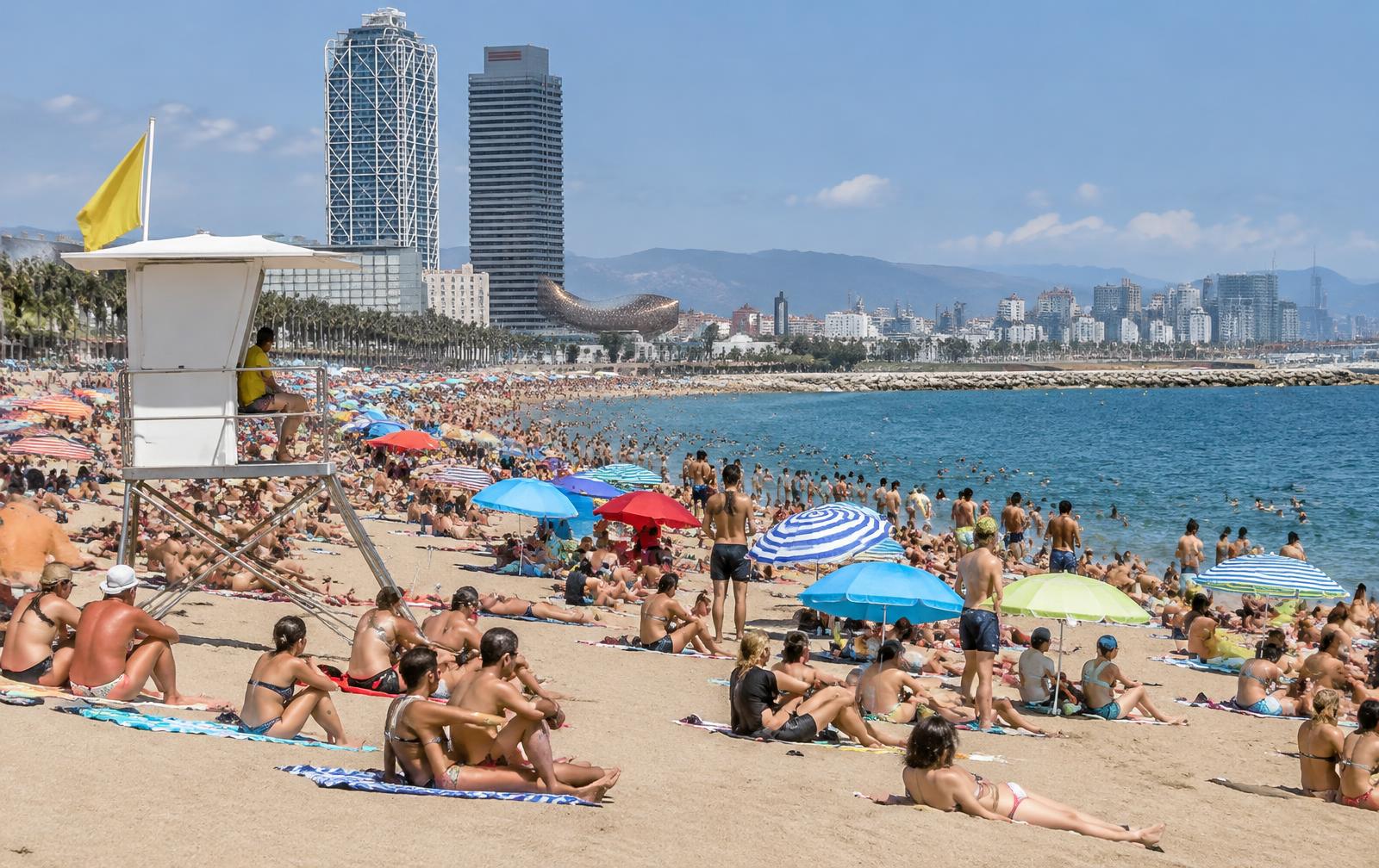 Symbolbild: Voller Stadtstrand in Barcelona mit Rettungsturm und Skyline (KI-generiert)